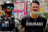 A handcuffed man wearing a black t shirt with "courage" printed across his chest squints as riot police move him.