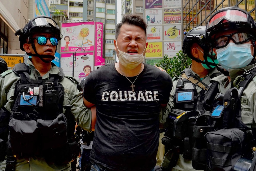 A handcuffed man wearing a black t shirt with "courage" printed across his chest squints as riot police move him.