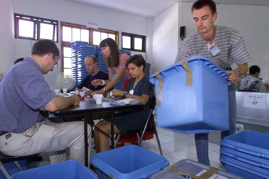 A man carries a blue box while people are casting their votes next to him.