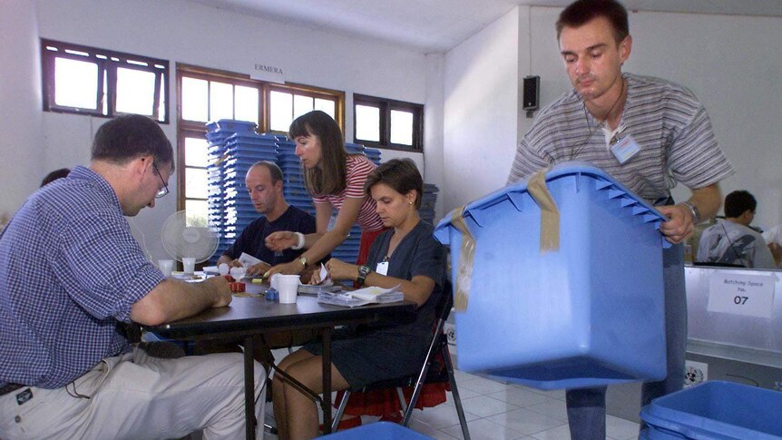A man carries a blue box while people are casting their votes next to him.