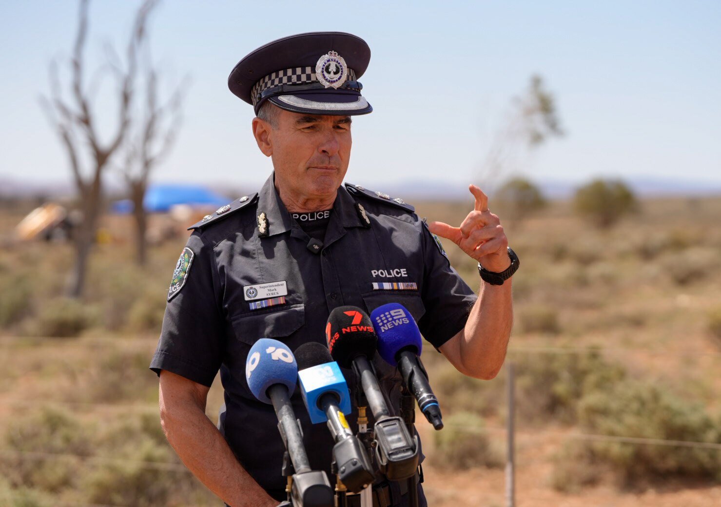 A police officer wears a uniform and hat. he has microphones in front of him and stands in outback landscape