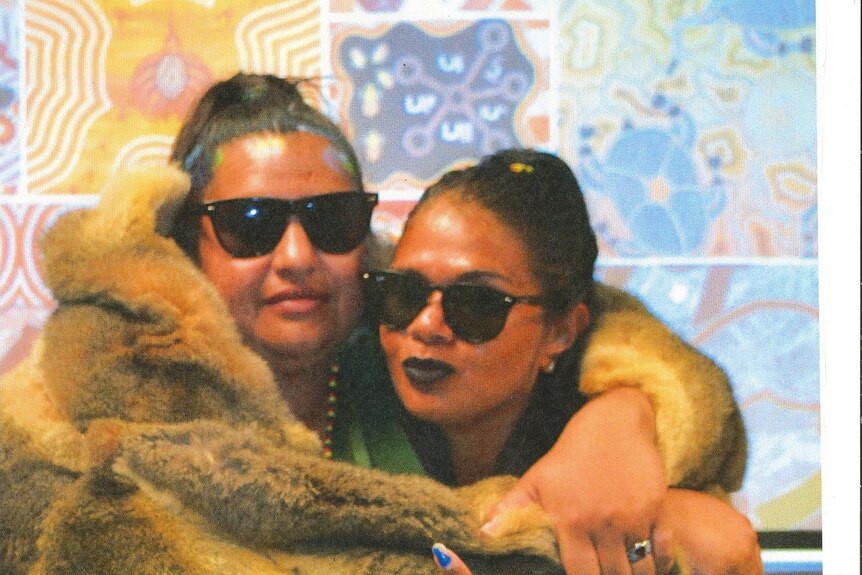 Heather and Suzzane both wear sunglasses as they hug, wrapped together in a possum-skin cloak.
