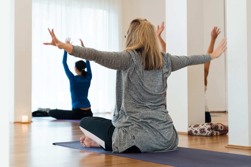 A woman sits on a yoga mat with her arms outstretched performing yoga in a studio.