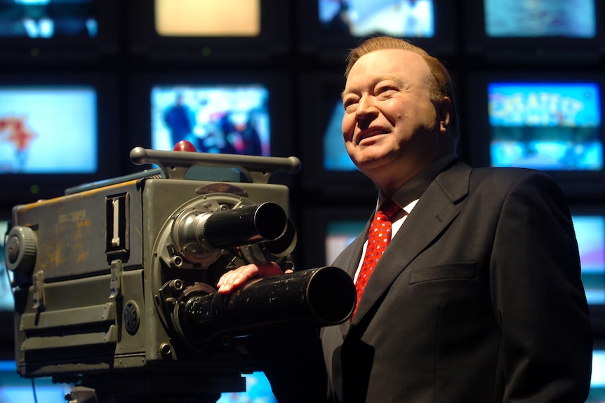 Bert Newton porte un costume et sourit à côté d'un appareil photo à l'ancienne avec des écrans de télévision derrière lui.