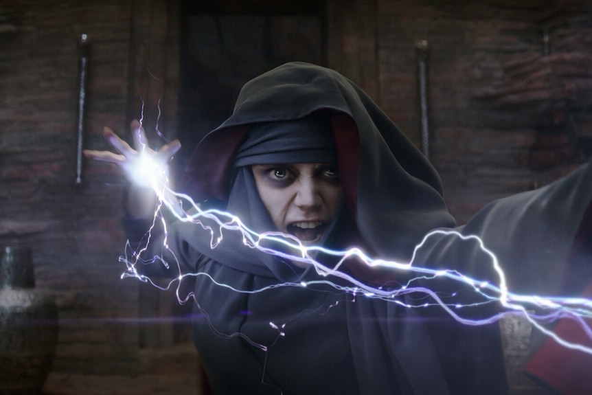 A young white girl with enlarged eyes wears a mystic's cloak and shoots a bolt of electricity from her hands in a darkened room.