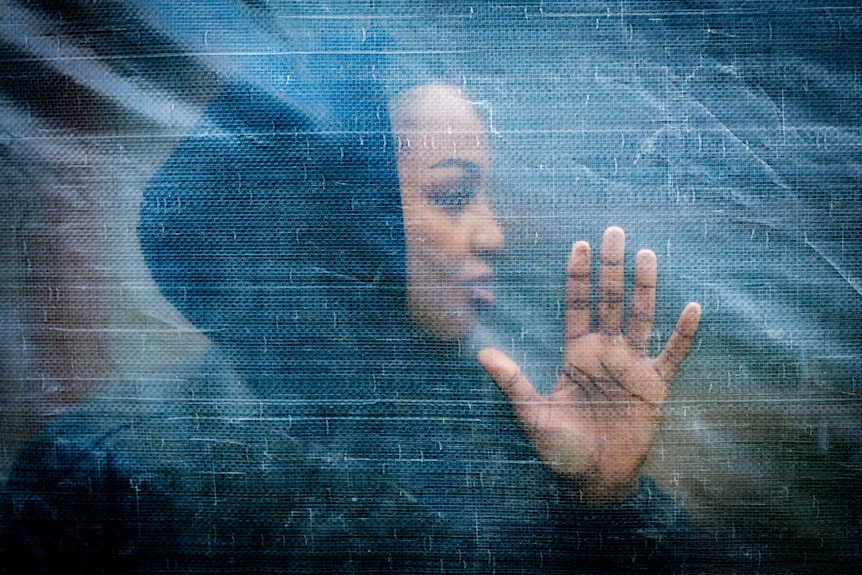 A young woman of African descent is seen through a plastic sheet with her hand pressed up against it