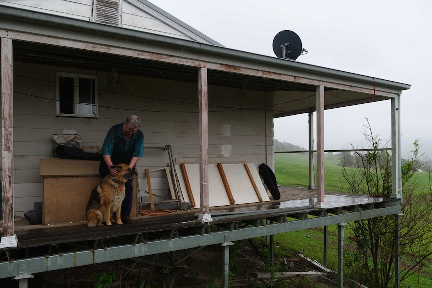 A man stands on the timber deck of a house with his dog, surrounded by green hills and paddocks.
