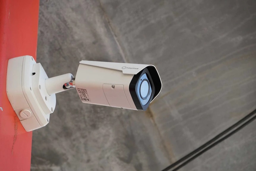 A rectangualr CCTV camera attached to a red post looking down 