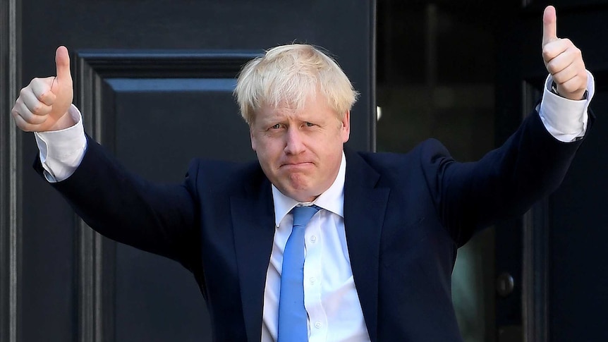 Boris Johnson wears a navy blue suit and light blue tie in front of a reflective black door.