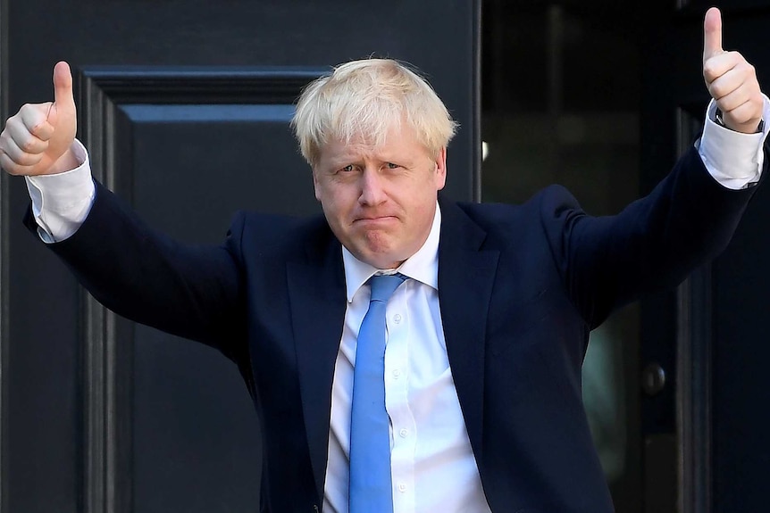 Boris Johnson wears a navy blue suit and light blue tie in front of a reflective black door.