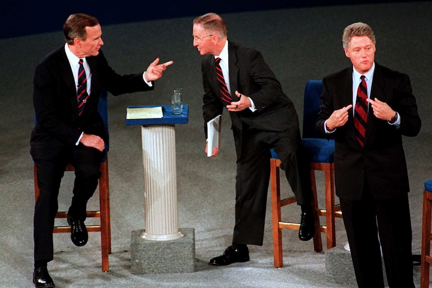 three men in suits on a stage on or near chairs