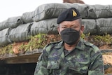 A man in camouflage gear, a black face mask and Royal Thai Army beret, sits ona a stack of sandbags 