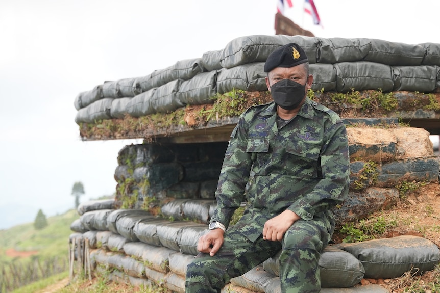 A man in camouflage gear, a black face mask and Royal Thai Army beret, sits ona a stack of sandbags 