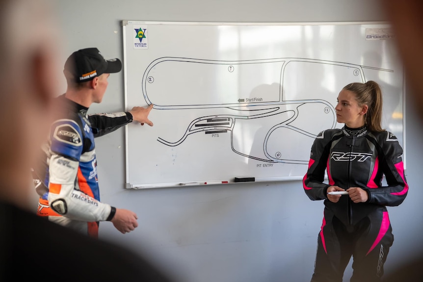 A woman and man stand in front of whiteboard with a motor track drawing, the man points to the track