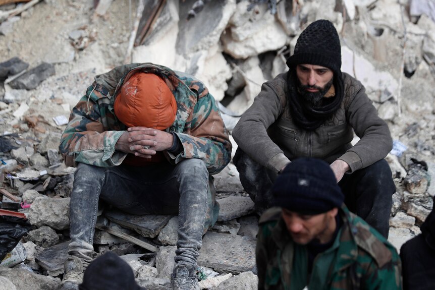 Two people sit among the rubble. One has their head in their hands, the other is staring sadly at the ground