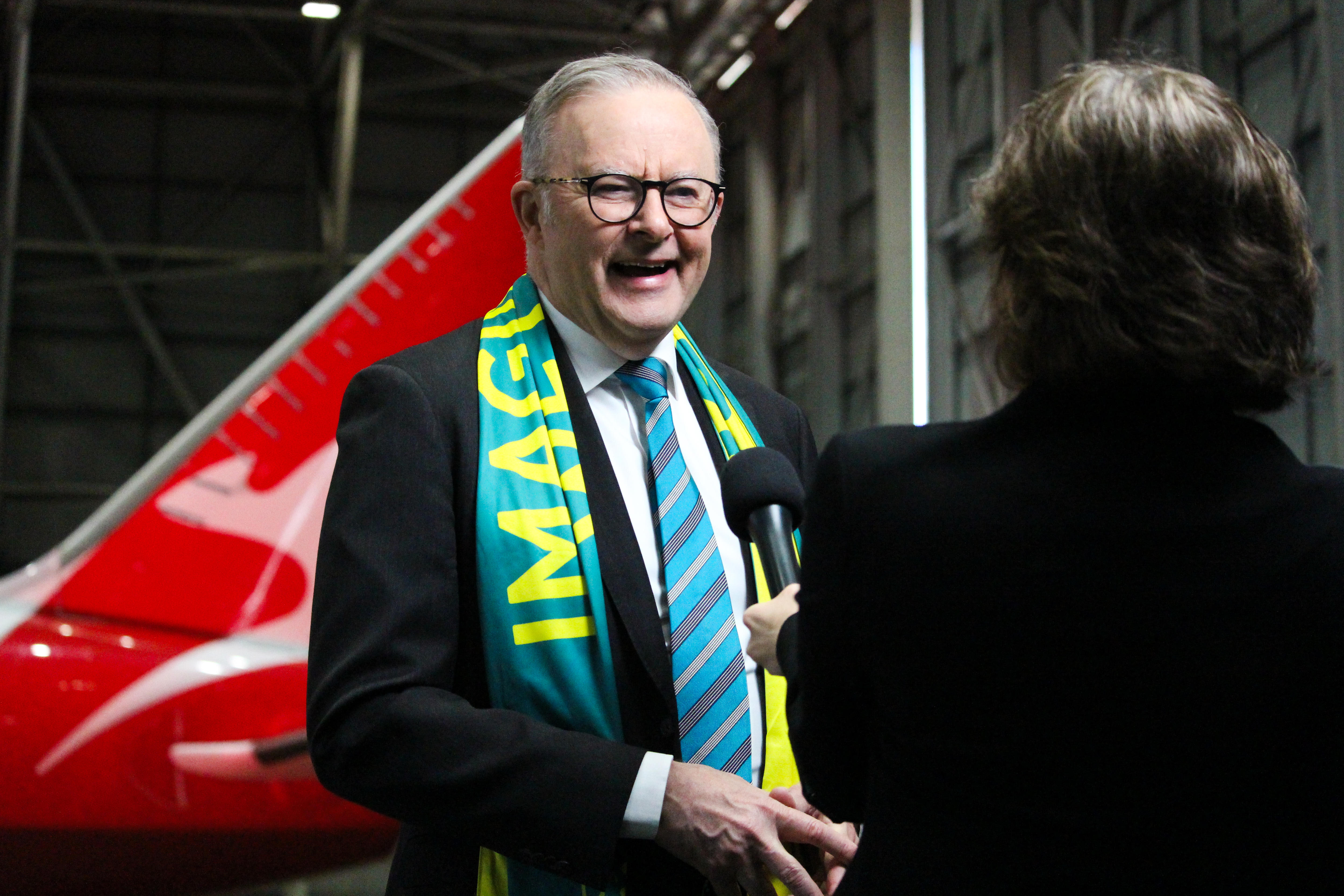 An older man with glasses wearing a green and gold scarf smiles, while being interviewed