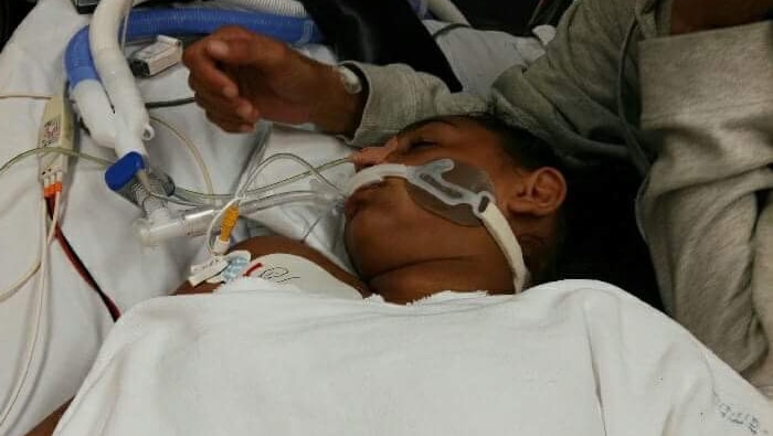 A girl lies on a hospital bed with tubes down her throat as her mother sits next to her