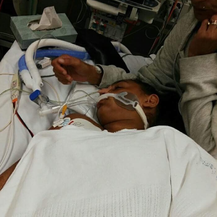 A girl lies on a hospital bed with tubes down her throat as her mother sits next to her