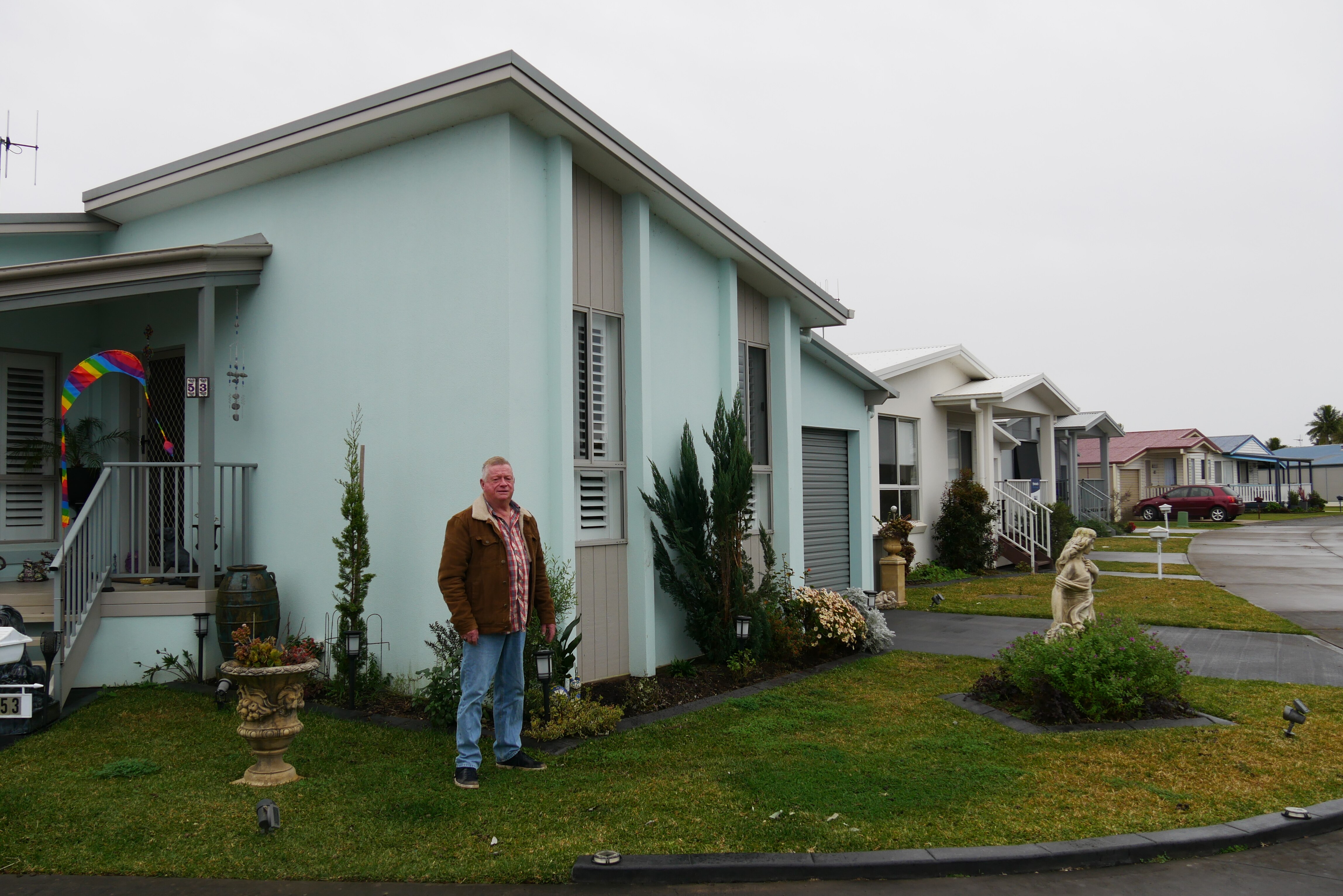 Man in a brown jacket stands in front of a blue house, surrounded by suburban neighborhood
