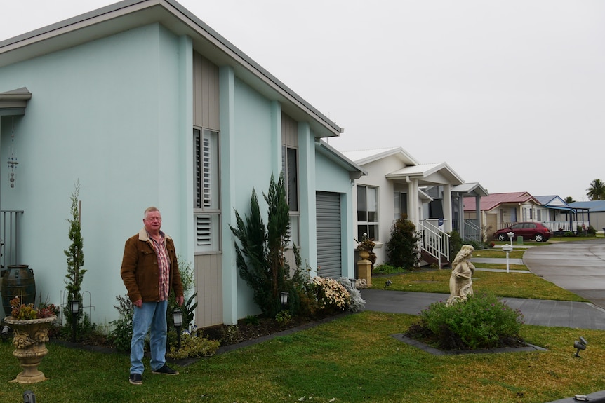 Man in a brown jacket stands in front of a blue house, surrounded by suburban neighborhood