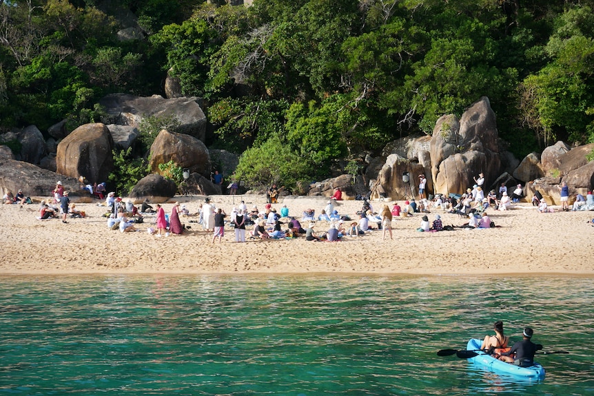 A crowd of people sit on a beach and watch a musician perform. Two people also watch from a kayak