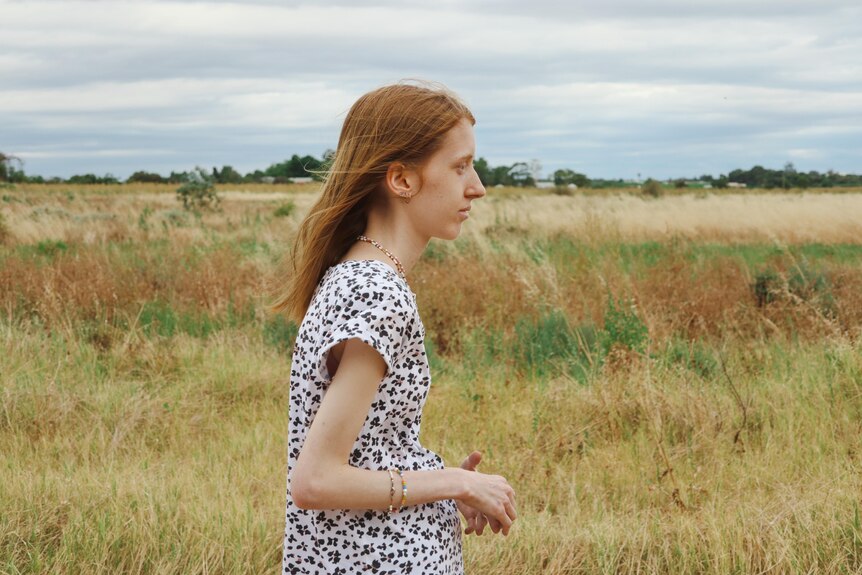 A young woman walks, behind her is a dry grass field, and a cloudy sky.