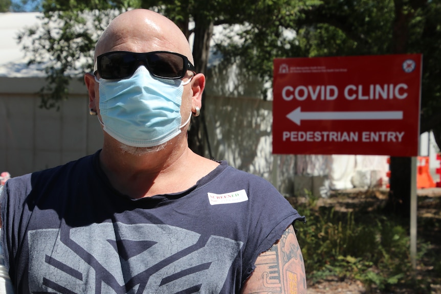 A strong looking bald man wears a sunglasses and mask pictured in front of a COVID clinic sign