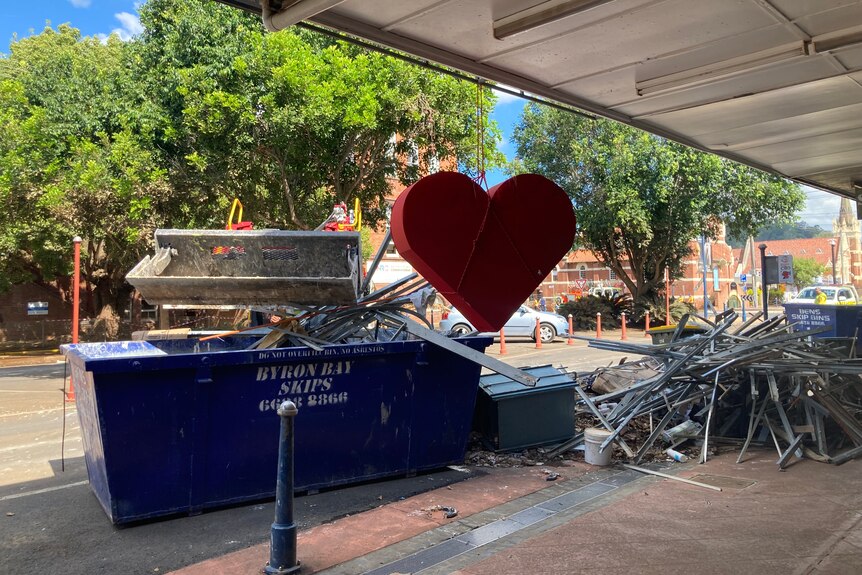 A large heart hangs from an outdoor roof near a skip bin and a pile of rubbish.