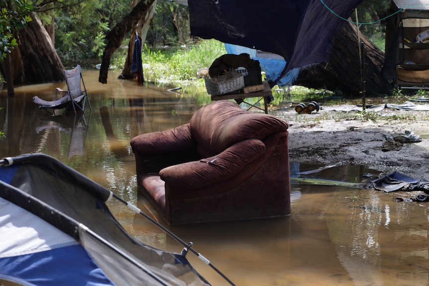 A brown velvet cough is half submerged in floodwater, with tents in the foreground and background also partially underwater.