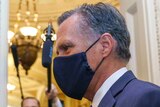 Senator Mitt Romney in profile wearing a mask as people stand around him with cameras and masks.