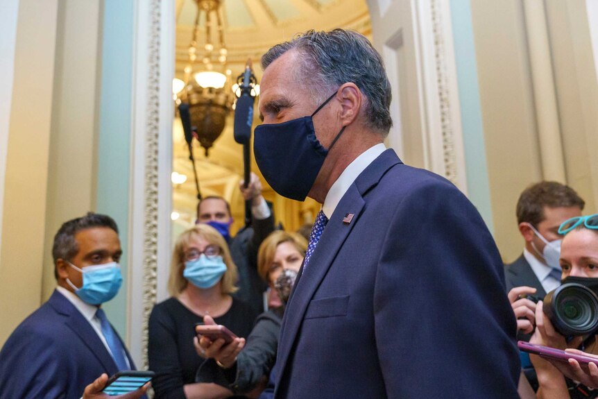 Senator Mitt Romney in profile wearing a mask as people stand around him with cameras and masks.