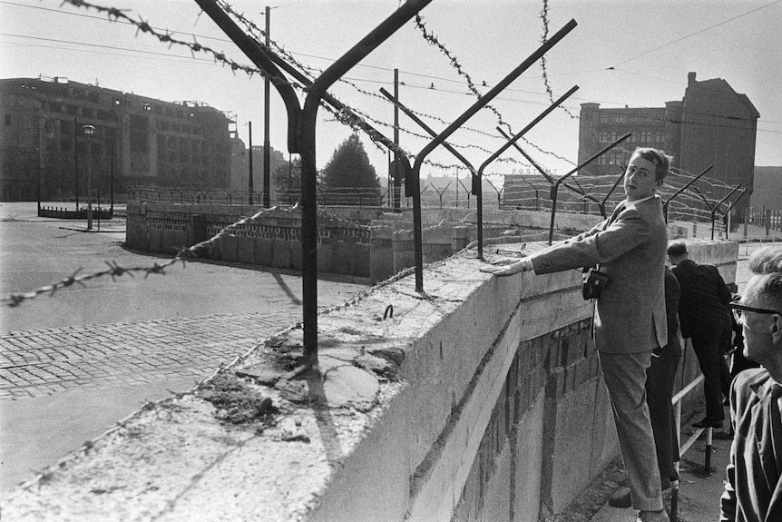 Men stand on a railing to look over the barbed wire-topped Berlin Wall, the deserted other side of the wall can be seen.