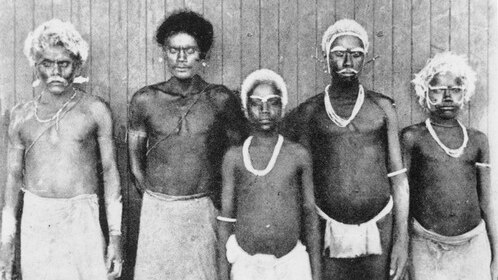 Five men in traditional islanders dress poses for a photo in front of a wooden wall