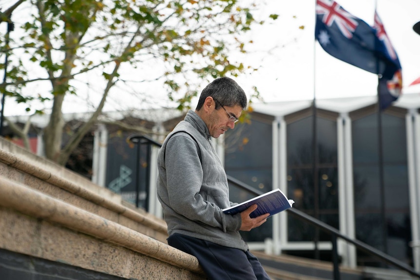 Mamutjan Abdurehim reads a book with the Australian flag floating in the background.