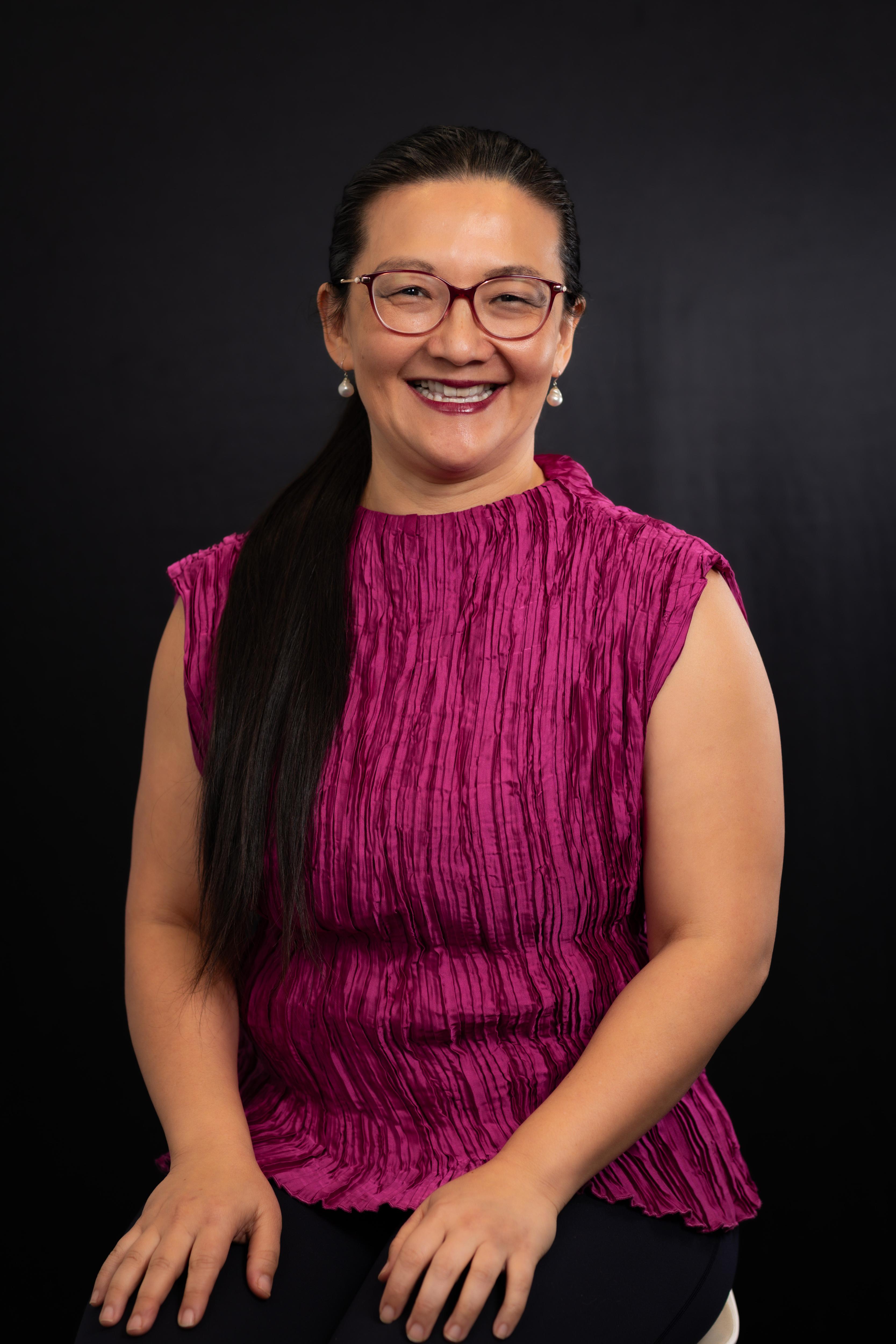 Una mujer con una camisa rosa oscuro, cabello largo y negro y gafas.