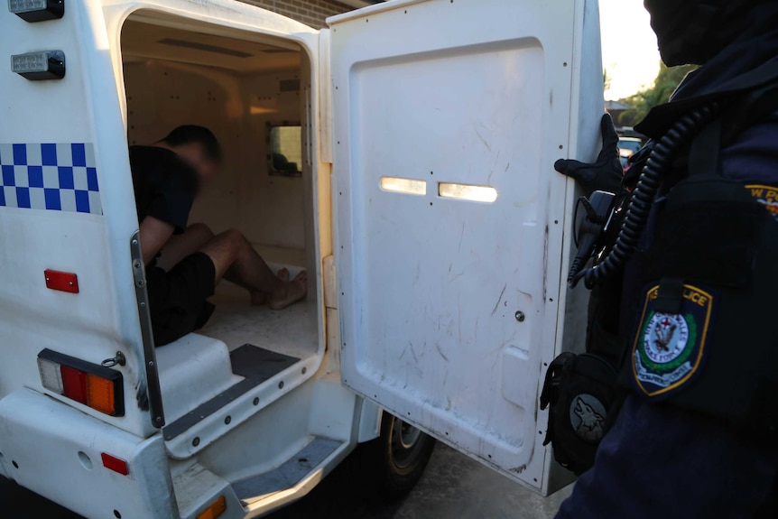 A police officer looks into the back of a police van, where a man is seated.