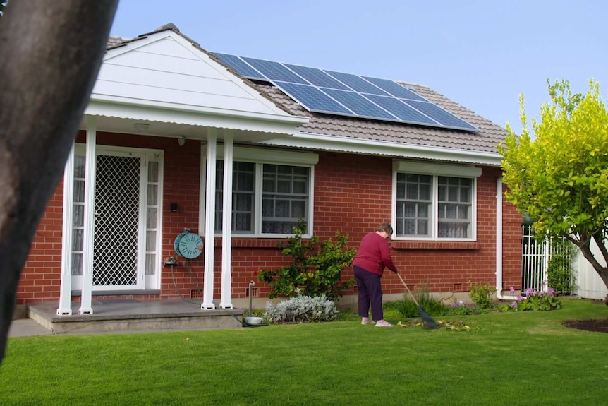 Marianne Hun rakes leaves in front of her red-brick house with solar panels on the roof.