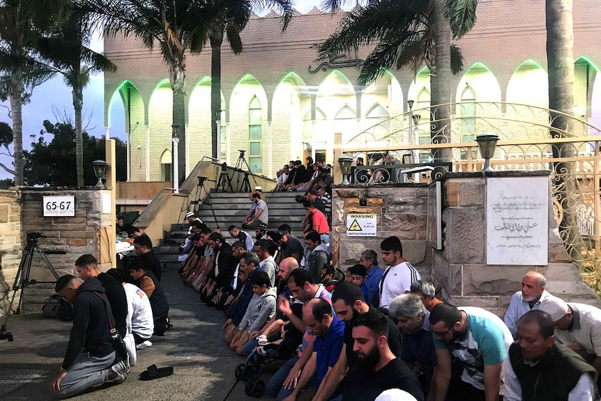 Lines of men kneel and place their hands on their knees as they face the same direction and pray outside a large mosque