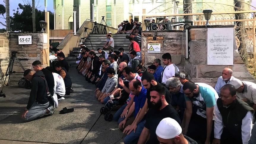 Lines of men kneel and place their hands on their knees as they face the same direction and pray outside a large mosque