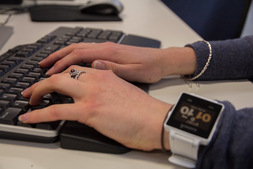 Close up of a woman's hands typing at a computer keyboard with some jewellery on and a large fitness watch on her wrist
