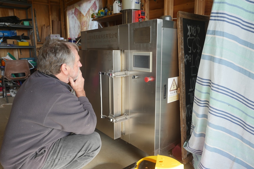 A man squats and scratches his chin while looking at a large stainless steel machine similar to an industrial oven.