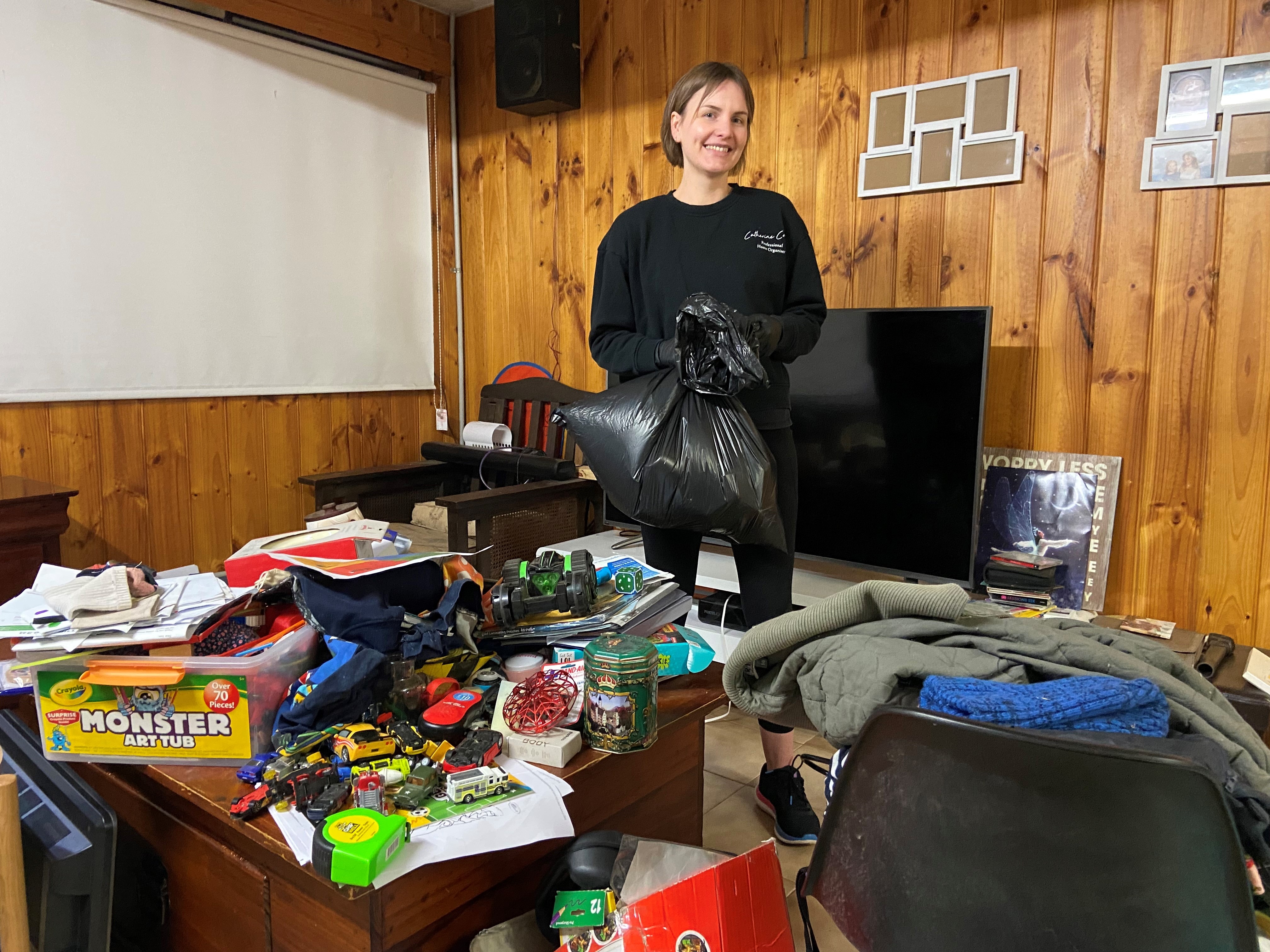 A woman stands in the middle of a TV room surrounded mess and piles of clothes.