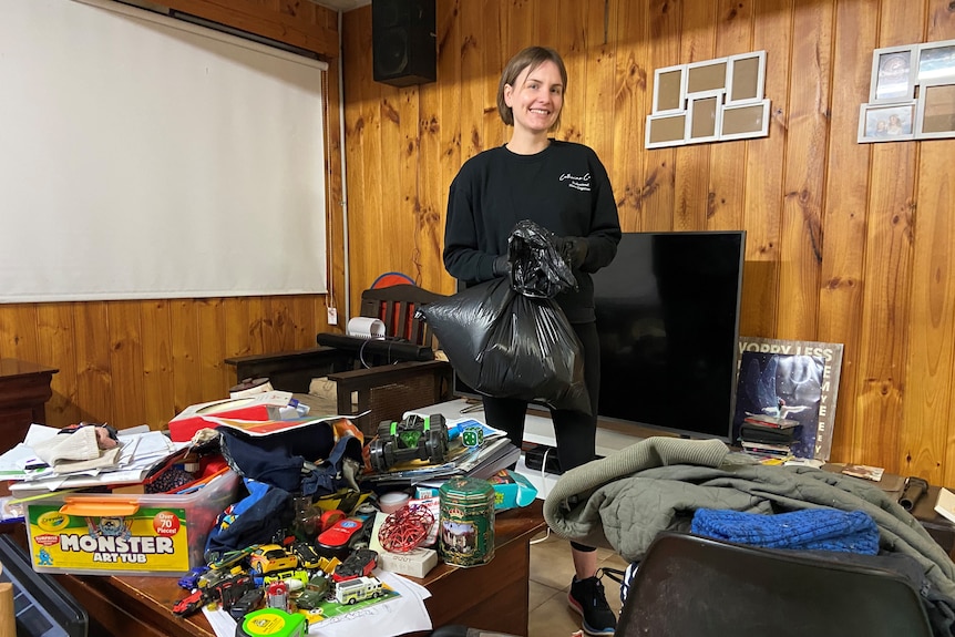 A woman stands in the middle of a TV room surrounded mess and piles of clothes.