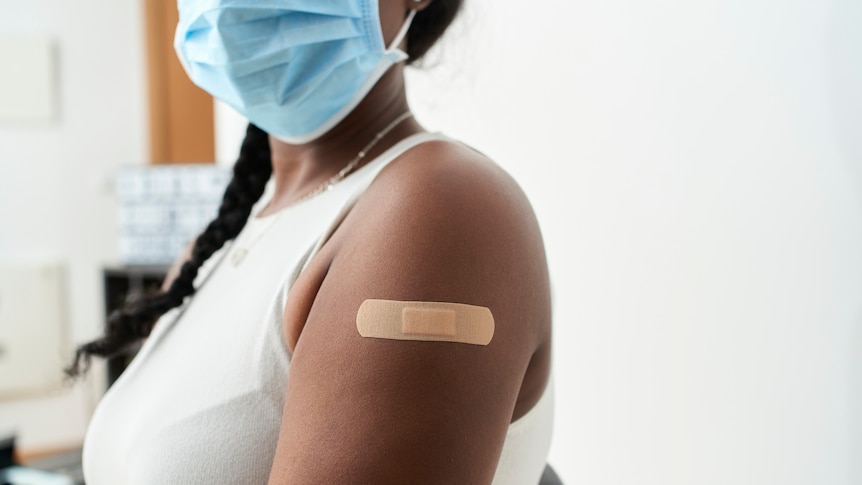 Woman with braid in her hair with white shirt and mask showing the band-aid on her arm after vaccination.