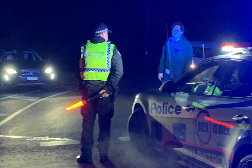 A police man holding lit up baton next to a police car talking to a man 