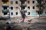 A man wearing a winter coat and beanie walks with his dog through the wreckage of an apartment building damaged by shelling.