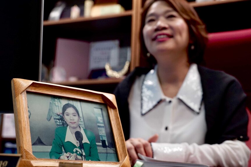 Priscilla Leung sits at a desk smiling. On the desk is a photo of her as a young student.