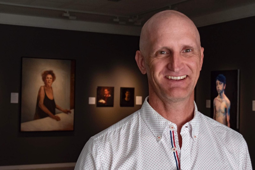 Man with shaved head stands in an art gallery with the portraits of a woman behind him on the left a young man on the right.