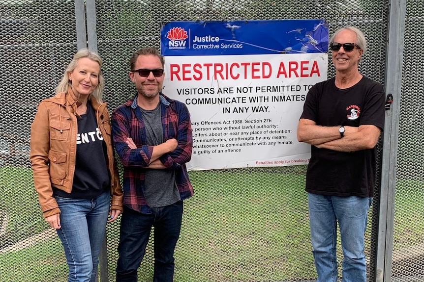 A woman and two men stand outside the fence of a prison, with a sign reading 'restricted area.'