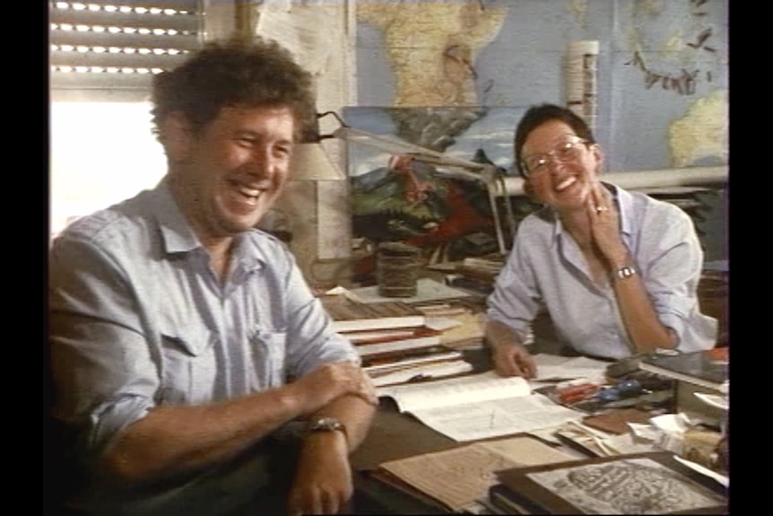 Middle-aged man with curly brown hair wears blue shirt and sit with white woman in similar shirt with glasses in home office.